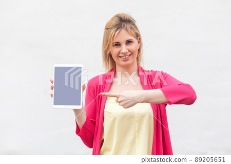 Portrait of satisfied beautiful young woman in pink blouse standing and holding tablet empty screen and pointing finger to device with toothy smile and looking at camera. Indoor, isolated, studio shot 89205651