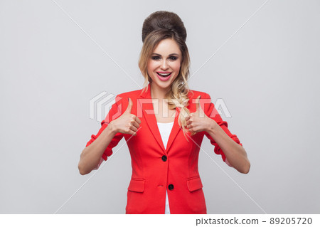 Portrait of glad beautiful business lady with hairstyle and makeup in red fancy blazer, standing, thumbs up and looking at camera with thumbs up. indoor studio shot, isolated on grey background. Portrait of glad beautiful business lady with hairstyle and makeup in red fancy blazer, standing, thumbs up and looking at camera with thumbs up. indoor studio shot, isolated on grey background. 89205720