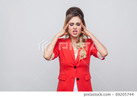 Portrait of sick beautiful business lady with hairstyle and makeup in red fancy blazer standing, holding her head and feeling headache. indoor studio shot, isolated on grey background. Portrait of sick beautiful business lady with hairstyle and makeup in red fancy blazer standing, holding her head and feeling headache. indoor studio shot, isolated on grey background. 89205744