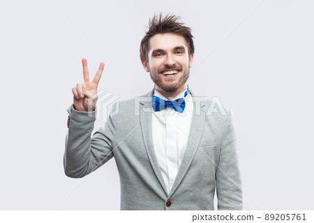 Portrait of happy toothy smiley handsome bearded man in casual grey suit, blue bow tie standing and looking at camera with victory or peace sign. indoor studio shot, isolated on light grey background. 89205761