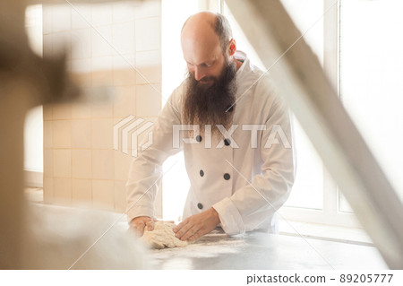 Adult baker with long beard in white uniform standing in his workplace and prepare the bread dough with hands kneading the dough on table with flour powder, Indoor, Concept of baking and patisserie Adult baker with long beard in white uniform standing in his workplace and prepare the bread dough with hands kneading the dough on table with flour powder, Indoor, Concept of baking and patisserie 89205777