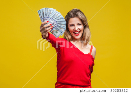 Portrait of excited rich beautiful woman in red dress standing showing dollar bills to camera, looking with open mouth, boasting salary or lottery win. indoor studio shot isolated on yellow background 89205812
