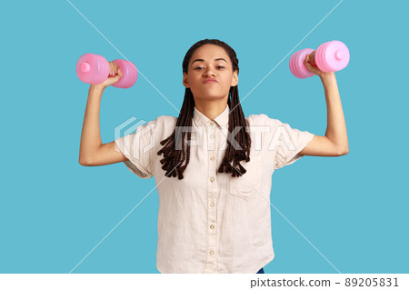 Assertive woman with black dreadlocks pumping up muscles holding dumbbells, looking at camera with confident expression, wearing white shirt. Indoor studio shot isolated on blue background. 89205831