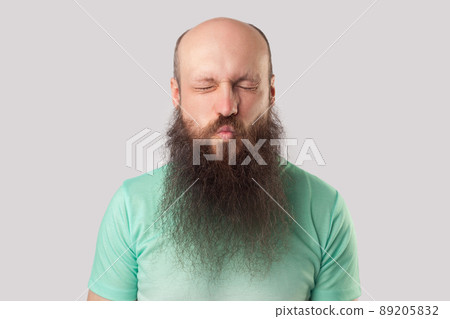 Portrait of funny middle aged bald man with long beard in light green t-shirt standing with closed eyes and kissing lips. indoor studio shot, isolated on grey background. Portrait of funny middle aged bald man with long beard in light green t-shirt standing with closed eyes and kissing lips. indoor studio shot, isolated on grey background. 89205832