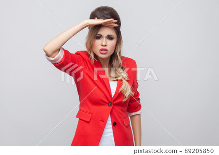 Portrait of attentive beautiful business lady with hairstyle and makeup in red fancy blazer, standing holding hand on forhead and looking far. indoor studio shot, isolated on grey background. Portrait of attentive beautiful business lady with hairstyle and makeup in red fancy blazer, standing holding hand on forhead and looking far. indoor studio shot, isolated on grey background. 89205850
