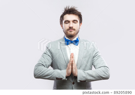 Portrait of calm relax handsome bearded man in casual grey suit and blue bow tie standing with palm hands, closed eyes and doing yoga meditating. indoor studio shot, isolated on light grey background. Portrait of calm relax handsome bearded man in casual grey suit and blue bow tie standing with palm hands, closed eyes and doing yoga meditating. indoor studio shot, isolated on light grey background. 89205890