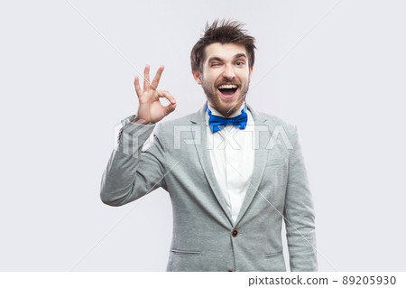 Portrait of funny happy handsome bearded man in casual grey suit and blue bow tie standing with Ok sign and looking at camera and winking. indoor studio shot, isolated on light grey background. Portrait of funny happy handsome bearded man in casual grey suit and blue bow tie standing with Ok sign and looking at camera and winking. indoor studio shot, isolated on light grey background. 89205930
