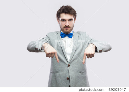 Here and right now. Portrait of serious bossy handsome bearded man in casual grey suit and blue bow tie standing pointing and looking at camera. indoor studio shot, isolated on light grey background. 89205947