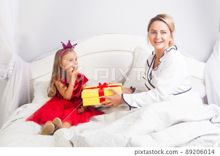 Amazing little girl in dress sitting in bed, looking surprised about gift box from mother, woman looking at camera with astonishment, greeting child on birthday morning, congratulation in bedroom Amazing little girl in dress sitting in bed, looking surprised about gift box from mother, woman looking at camera with astonishment, greeting child on birthday morning, congratulation in bedroom 89206014