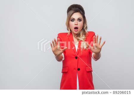 Portrait of shocked beautiful business lady with hairstyle and makeup in red fancy blazer, standing with blocking hands and looking at camera. indoor studio shot, isolated on grey background. Portrait of shocked beautiful business lady with hairstyle and makeup in red fancy blazer, standing with blocking hands and looking at camera. indoor studio shot, isolated on grey background. 89206015