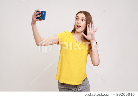 Portrait of happy curly-haired teen girl in yellow T-shirt taking selfie, having conversation on video call and waving hello gesture, vlog broadcast. Indoor studio shot isolated on gray background. 89206016