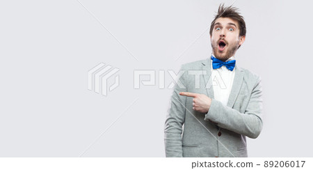 Portrait of amazed handsome bearded man in casual grey suit and blue bow tie standing looking at camera with surprised face and pointing at copyspace. studio shot, isolated on light grey background. Portrait of amazed handsome bearded man in casual grey suit and blue bow tie standing looking at camera with surprised face and pointing at copyspace. studio shot, isolated on light grey background. 89206017