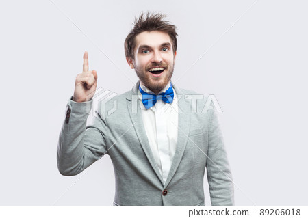 have Idea. Portrait of handsome excited bearded man in casual grey suit and blue bow tie standing and looking at camera with toothy smile. indoor studio shot, isolated on light grey background. 89206018