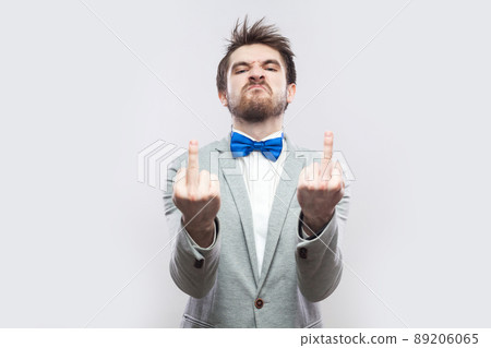 Portrait of angry handsome bearded man in casual grey suit and blue bow tie standing with middle finger fuck sing and looking at camera. indoor studio shot, isolated on light grey background. Portrait of angry handsome bearded man in casual grey suit and blue bow tie standing with middle finger fuck sing and looking at camera. indoor studio shot, isolated on light grey background. 89206065
