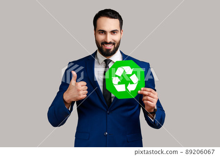 Portrait of satisfied delighted bearded businessman holding pointing at green recycling sign, saving environment, wearing official style suit. Indoor studio shot isolated on gray background. Portrait of satisfied delighted bearded businessman holding pointing at green recycling sign, saving environment, wearing official style suit. Indoor studio shot isolated on gray background. 89206067