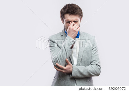 Portrait of sad alone depressed handsome bearded man in casual grey suit and blue bow tie standing, holding head down and crying. indoor studio shot, isolated on light grey background. 89206072