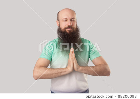 Portrait of calm serious middle aged bald man with long beard in light green t-shirt standing with closed eyes, palm hand and doing yoga meditation. indoor studio shot, isolated on grey background. Portrait of calm serious middle aged bald man with long beard in light green t-shirt standing with closed eyes, palm hand and doing yoga meditation. indoor studio shot, isolated on grey background. 89206098