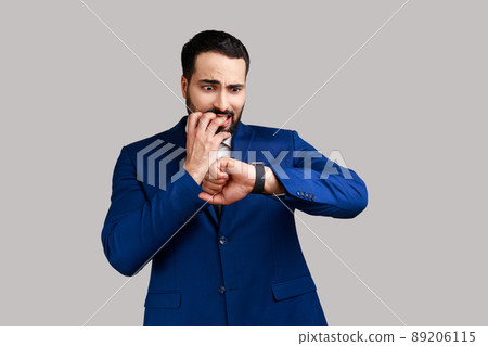 Nervous bearded man biting finger nails and looking at his wrist clock with anxious, worried about deadline, wearing official style suit. Indoor studio shot isolated on gray background. Nervous bearded man biting finger nails and looking at his wrist clock with anxious, worried about deadline, wearing official style suit. Indoor studio shot isolated on gray background. 89206115