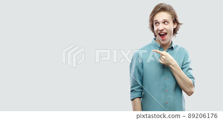 Portrait of amazed handsome long haired blonde young man in blue casual shirt standing and pointing at side copyspace with surprised funny face. indoor studio shot, isolated on light grey background. Portrait of amazed handsome long haired blonde young man in blue casual shirt standing and pointing at side copyspace with surprised funny face. indoor studio shot, isolated on light grey background. 89206176