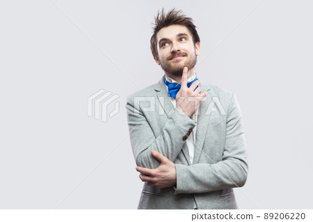 Portrait of dreamful happy handsome bearded man in casual grey suit and blue bow tie standing and looking away, smiling and dreaming. indoor studio shot, isolated on light grey background. Portrait of dreamful happy handsome bearded man in casual grey suit and blue bow tie standing and looking away, smiling and dreaming. indoor studio shot, isolated on light grey background. 89206220