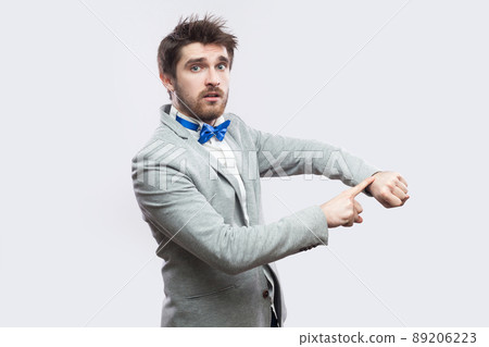 Portrait of serious worried handsome bearded man in casual grey suit and blue bow tie standing with watch hand gesture and looking at camera. indoor studio shot, isolated on light grey background. 89206223
