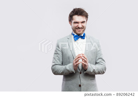 Portrait of cunning handsome bearded man in casual grey suit and blue bow tie standing with funny face and palm hand looking at camera with idea. indoor studio shot, isolated on light grey background. Portrait of cunning handsome bearded man in casual grey suit and blue bow tie standing with funny face and palm hand looking at camera with idea. indoor studio shot, isolated on light grey background. 89206242