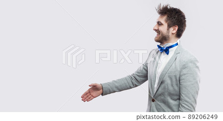 Profile side view portrait of happy handsome bearded man in casual grey suit, blue bow tie standing and looking straight with toothy smile and greeting. studio shot, isolated on light grey background. Profile side view portrait of happy handsome bearded man in casual grey suit, blue bow tie standing and looking straight with toothy smile and greeting. studio shot, isolated on light grey background. 89206249