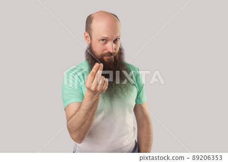 Portrait of funny middle aged bald man with long beard in light green t-shirt standing with money or italian gesture and looking at camera. indoor studio shot, isolated on grey background. 89206353
