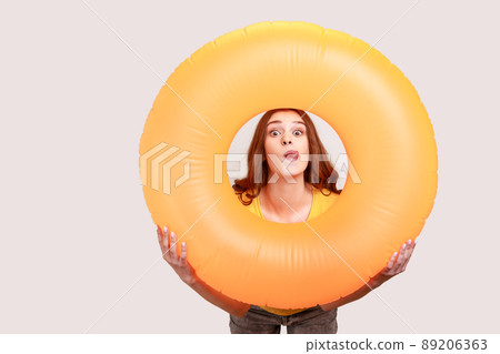 Teen girl of with brown wavy hair in yellow T-shirt holding orange rubber ring, having fun on resort, looking at camera and showing tongue out. Indoor studio shot isolated on gray background. Teen girl of with brown wavy hair in yellow T-shirt holding orange rubber ring, having fun on resort, looking at camera and showing tongue out. Indoor studio shot isolated on gray background. 89206363