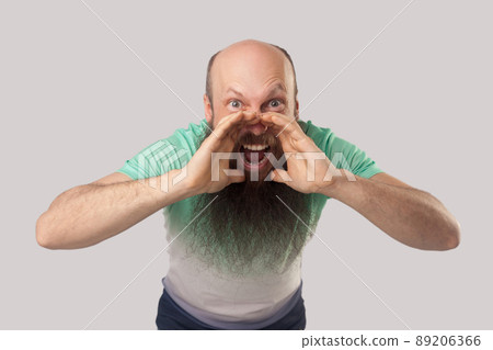 Portrait of middle aged bald man with long beard in light green t-shirt standing looking and screaming with raised arms. indoor studio shot, isolated on grey background. Portrait of middle aged bald man with long beard in light green t-shirt standing looking and screaming with raised arms. indoor studio shot, isolated on grey background. 89206366