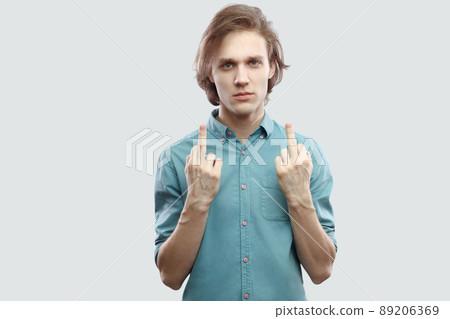 Portrait of poker face handsome long haired blonde young man in blue casual shirt standing and looking at camera with middle finger fuck sign. indoor studio shot, isolated on light grey background. 89206369