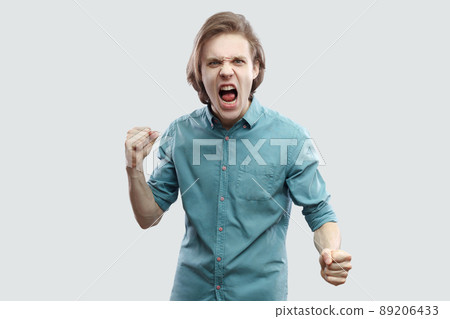 Portrait of angry crazy handsome long haired blonde young man in blue casual shirt standing, looking at camera, screaming and ready to attack. indoor studio shot, isolated on light grey background. 89206433