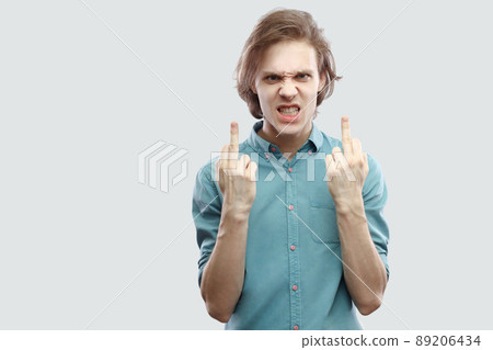 Portrait of angry handsome long haired blonde young man in blue casual shirt standing and looking at camera with middle finger fuck sign. indoor studio shot, isolated on light grey background. 89206434