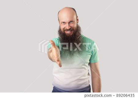 Portrait of happy middle aged bald man with long beard in green t-shirt standing, looking with toothy smile, giving hand to handshake and greeting. indoor studio shot, isolated on grey background. 89206435