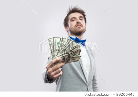 Take it! Portrait of confident attractive young businessman in gray coat and blue bow tie standing, proudly with head up and giving you many dollars. Indoor, studio shot, isolated, gray background. 89206445