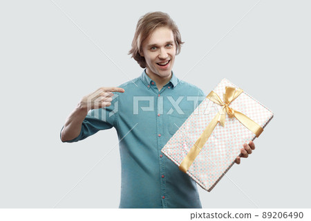 It`s yours! Satisfied modern young man in light blue shirt standing and pointing finger to present with yellow bow and toothy smile, looking at camera. Indoor, isolated, studio shot, grey background 89206490