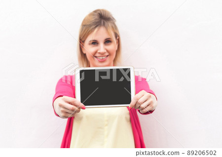 Portrait of happy beautiful young businesswoman in pink blouse standing and holding tablet empty screen with toothy smile and looking at camera. Indoor, isolated, copy space, white background 89206502
