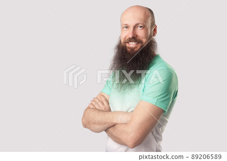 Portrait of happy satisfied middle aged bald man with long beard in green t-shirt standing with crossed arms and looking at camera with toothy smile. indoor studio shot, isolated on grey background. 89206589