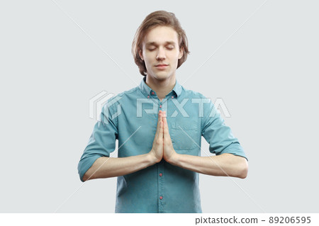 Portrait of calm handsome long haired blonde young man in blue casual shirt standing in yoga pose and meditating, and try to relax. indoor studio shot, isolated on light grey background. Portrait of calm handsome long haired blonde young man in blue casual shirt standing in yoga pose and meditating, and try to relax. indoor studio shot, isolated on light grey background. 89206595