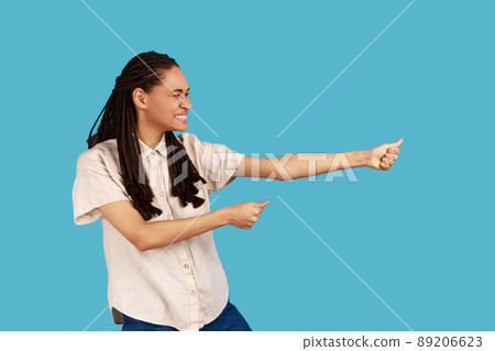Profile portrait strong assertive woman with dreadlocks pulling invisible rope, showing her persistence and leadership qualities, wearing white shirt. Indoor studio shot isolated on blue background. 89206623