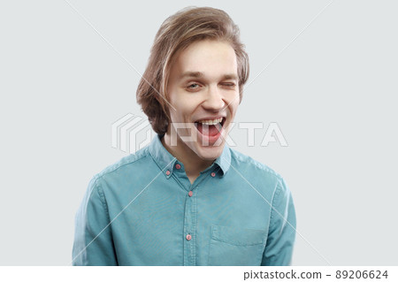 Portrait of funny happy handsome long haired blonde young man in blue casual shirt standing and looking at camera winking. indoor studio shot, isolated on light grey background. 89206624