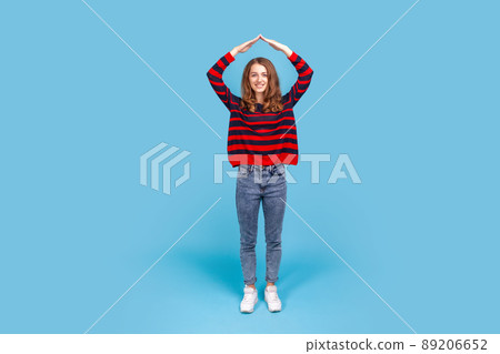 Full length portrait of smiling friendly woman, doing house roof gesture over head, feeling herself in safety, wearing striped casual style sweater. Indoor studio shot isolated on blue background. 89206652