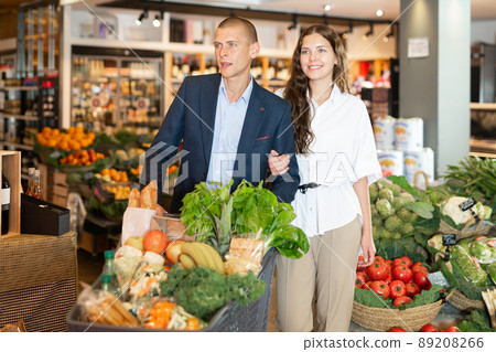 Happy couple is standing with trolley with food for dinner in supermarket Happy couple is standing with trolley with food for dinner in supermarket 89208266