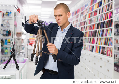 Buyer carefully chooses a tripod for camera in an electronics store 89208481