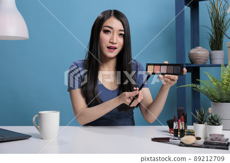 Asian woman influencer holding palette showing at camera while doing makeup tutorial advertising beauty product. Social media content creator recording cosmetic review for vlogging channel Asian woman influencer holding palette showing at camera while doing makeup tutorial advertising beauty product. Social media content creator recording cosmetic review for vlogging channel 89212709