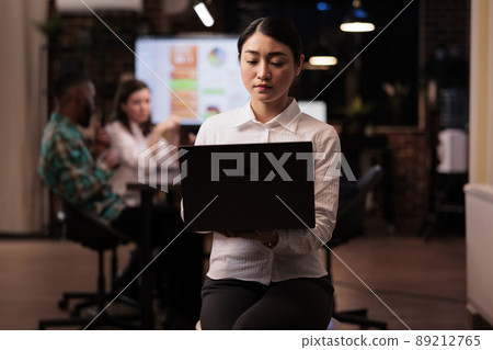 Portrait of asian woman holding laptop in startup office during late night meeting looking at screen. Employee posing confident working overtime with mixed team brainstorming marketing strategy. Portrait of asian woman holding laptop in startup office during late night meeting looking at screen. Employee posing confident working overtime with mixed team brainstorming marketing strategy. 89212765