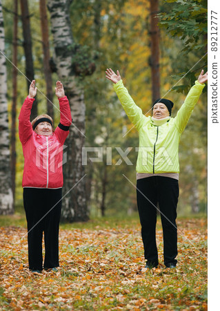 Elderly women perform a warm-up and raise their hands up in the autumn park after a scandinavian walk 89212777