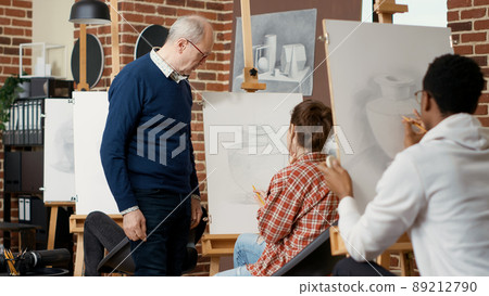 Elder teacher guiding young student to draw vase sketch on canvas, attending art program to develop creativity skills and find inspiration. Artwork drawing using artistic tools. Tripod shot Elder teacher guiding young student to draw vase sketch on canvas, attending art program to develop creativity skills and find inspiration. Artwork drawing using artistic tools. Tripod shot 89212790