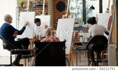 Old woman with disability using pencil to draw vase artwork, attending creative lesson in wheelchair to develop new drawing skills. Educational activity in workshop. Tripod shot. Old woman with disability using pencil to draw vase artwork, attending creative lesson in wheelchair to develop new drawing skills. Educational activity in workshop. Tripod shot. 89212791