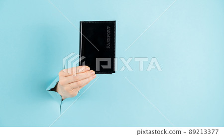 A woman's hand sticks out of a hole in a blue paper background and holds a passport in a black cover. A woman's hand sticks out of a hole in a blue paper background and holds a passport in a black cover. 89213377